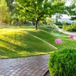 Sistema automatico di irrigazione del giardino con diversi irrigatori installati sotto il manto erboso. Progettazione paesaggistica con colline a prato e frutteto irrigati con irroratrici autonome intelligenti al tramonto della sera