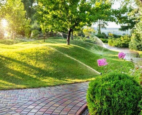 Sistema automatico di irrigazione del giardino con diversi irrigatori installati sotto il manto erboso. Progettazione paesaggistica con colline a prato e frutteto irrigati con irroratrici autonome intelligenti al tramonto della sera